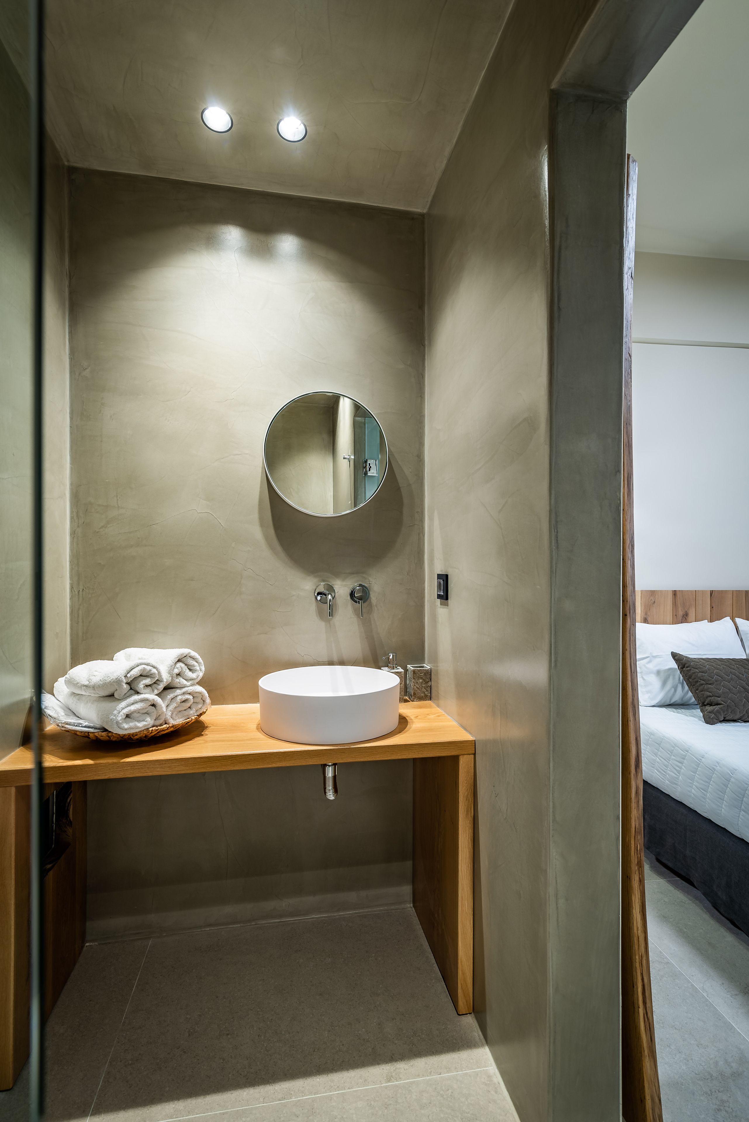 Un baño moderno con un espejo redondo, un tocador de madera y una cama visible a través de una puerta abierta.