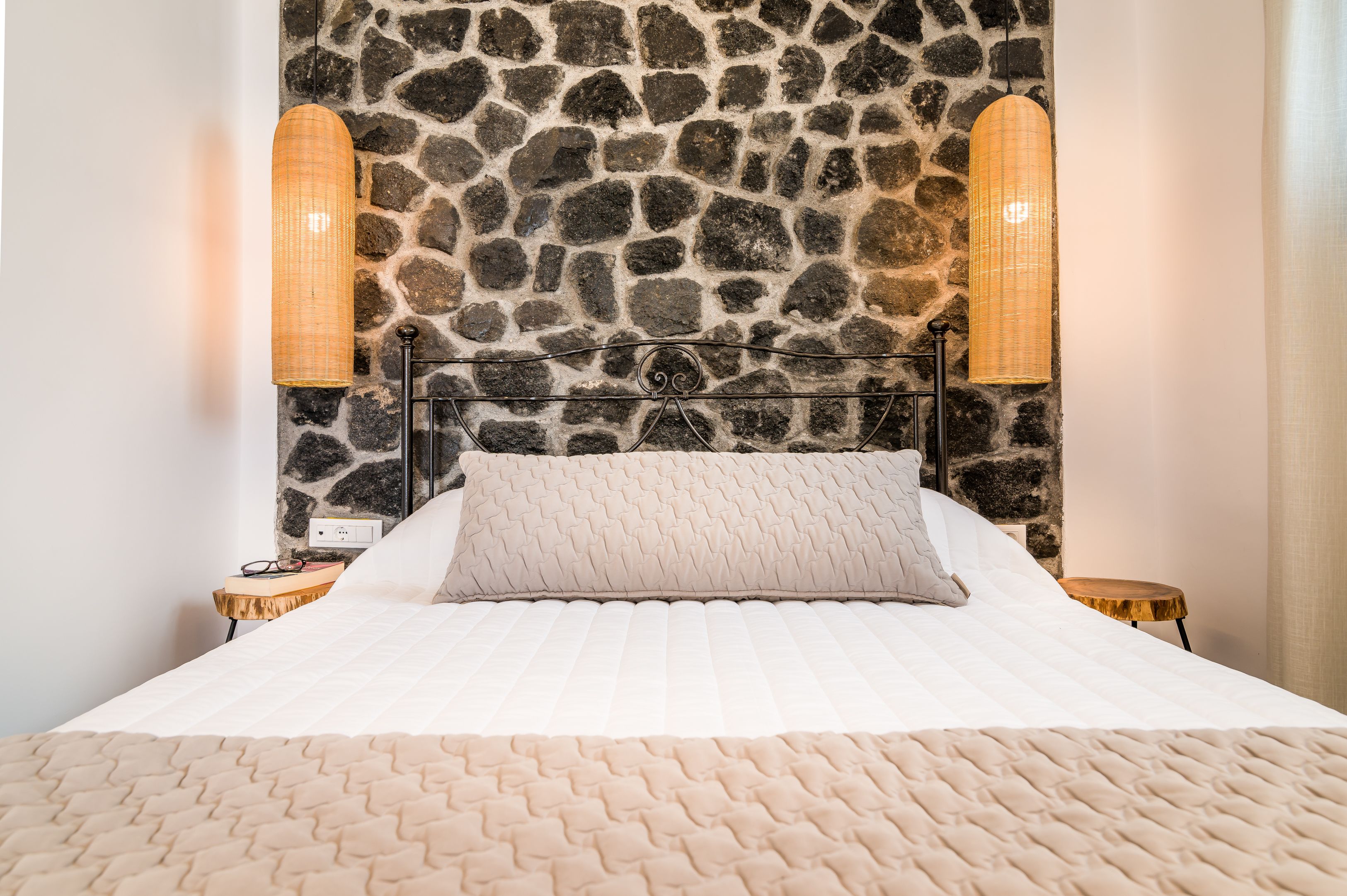Ein Bett mit einer Steinwand dahinter, zwei Nachttischlampen und ein Nachttisch mit einem Brillenpaar.