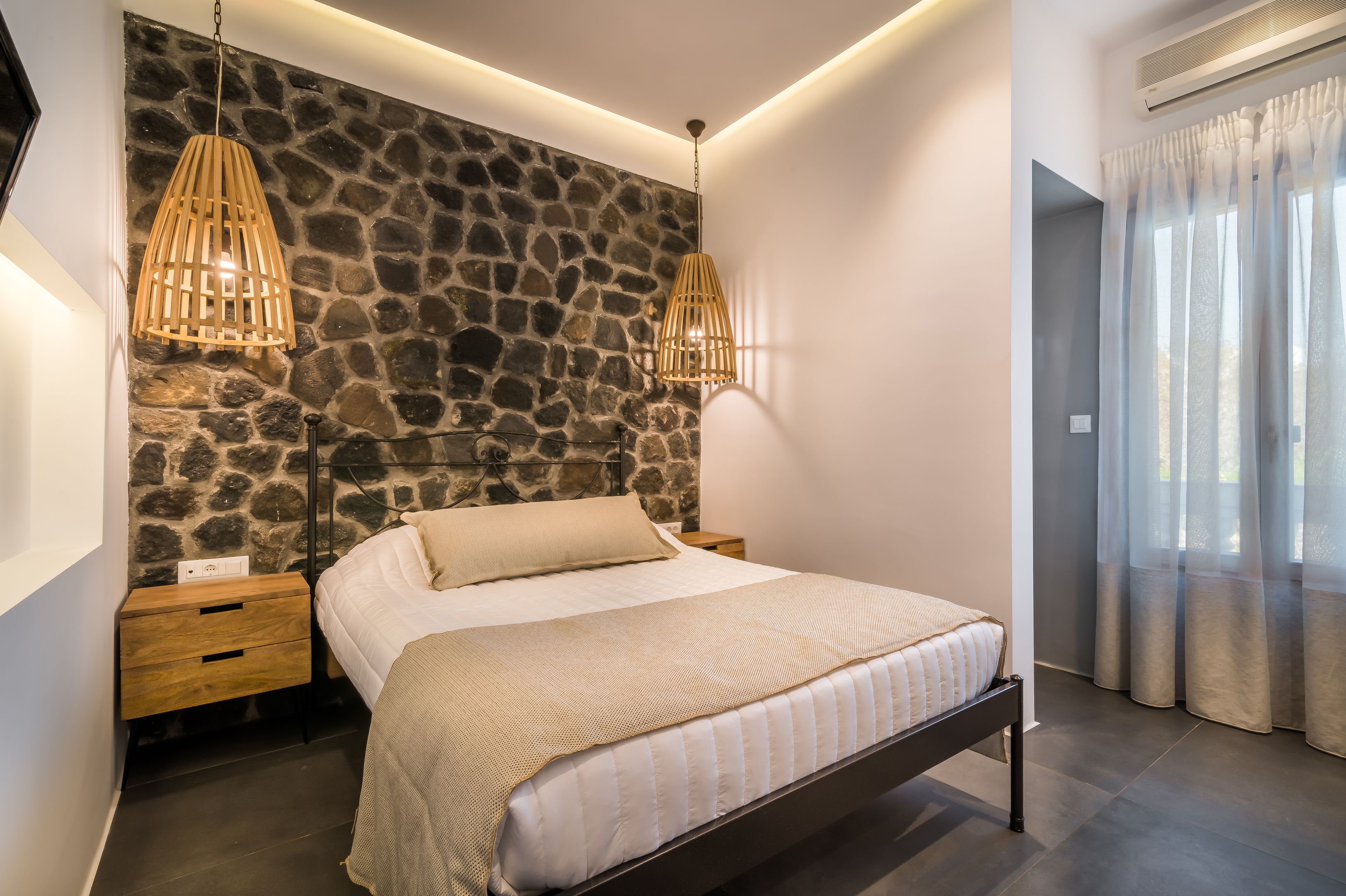 Una camera da letto con parete in pietra, un letto con copriletto beige, comodini in legno e due lampade a sospensione.