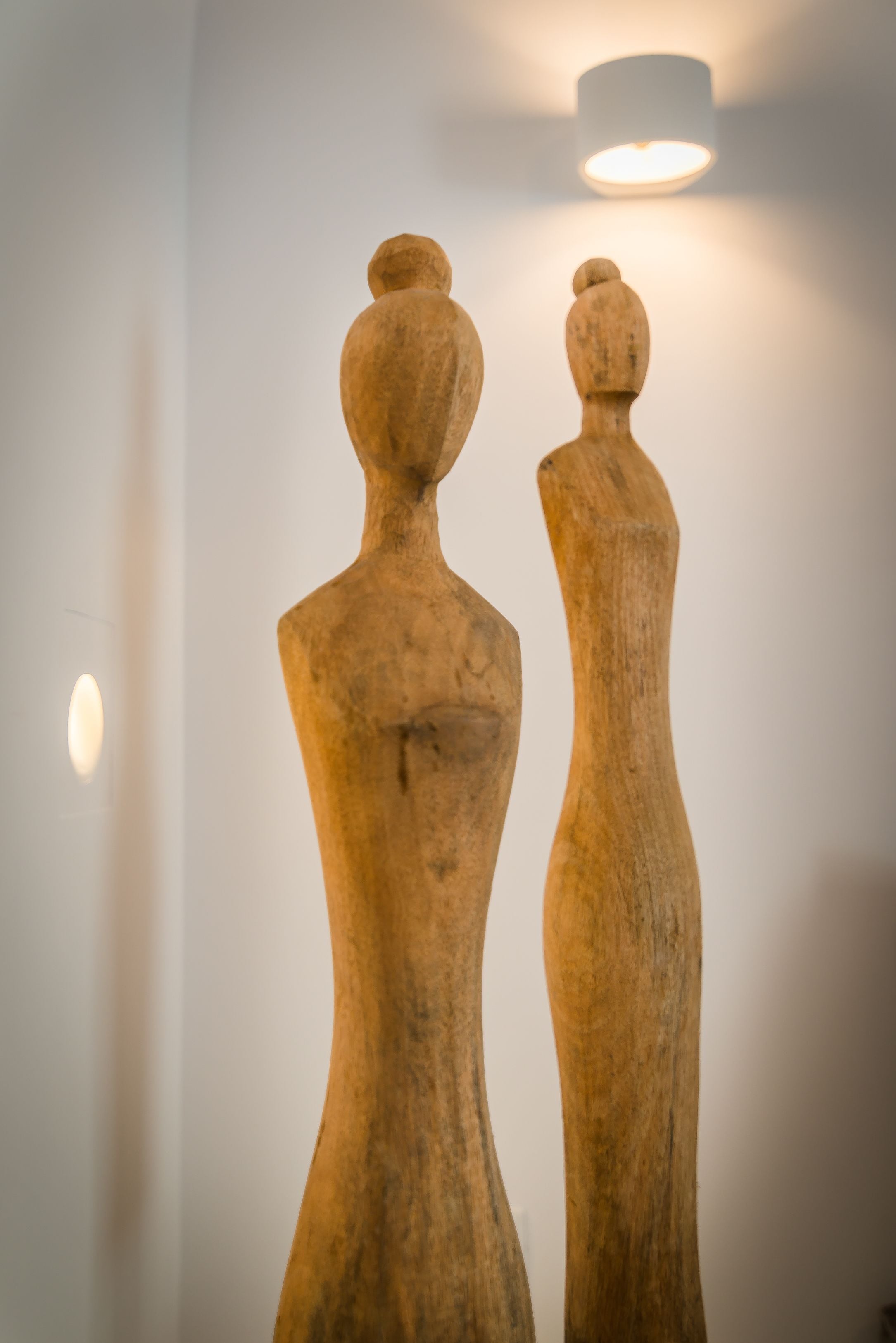 Two wooden sculptures against a plain wall with a ceiling light fixture.