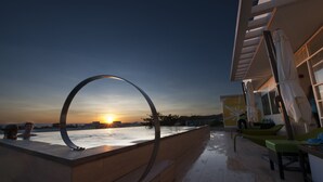 Een zwembad zonder rand met uitzicht op een zonsondergang, een modern architectonisch ontwerp, en een loungegedeelte met stoelen en parasols.