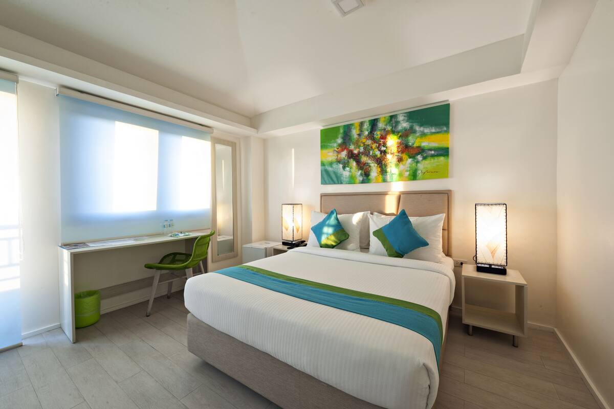 Ein Hotelzimmer mit einem großen Bett, einem Schreibtisch und einem Sessel. An der Wand hängt ein Bild und es gibt zwei Nachttischlampen.