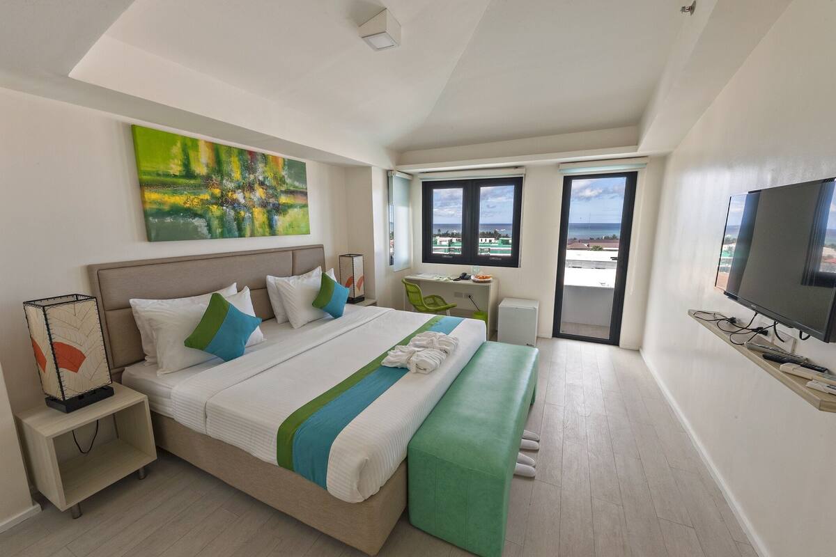Ein modernes Hotelzimmer mit einem großen Bett, einem Schreibtisch, einem Sessel, einem Fernseher und einem Balkon mit Blick.