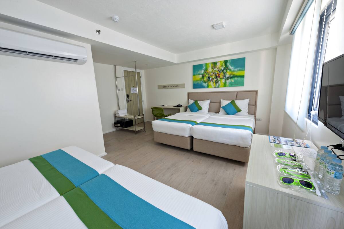 Ein Hotelzimmer mit zwei Betten, einem Schreibtisch, einem Stuhl, einer Klimaanlage, einem Fernseher und einem Bild an der Wand.