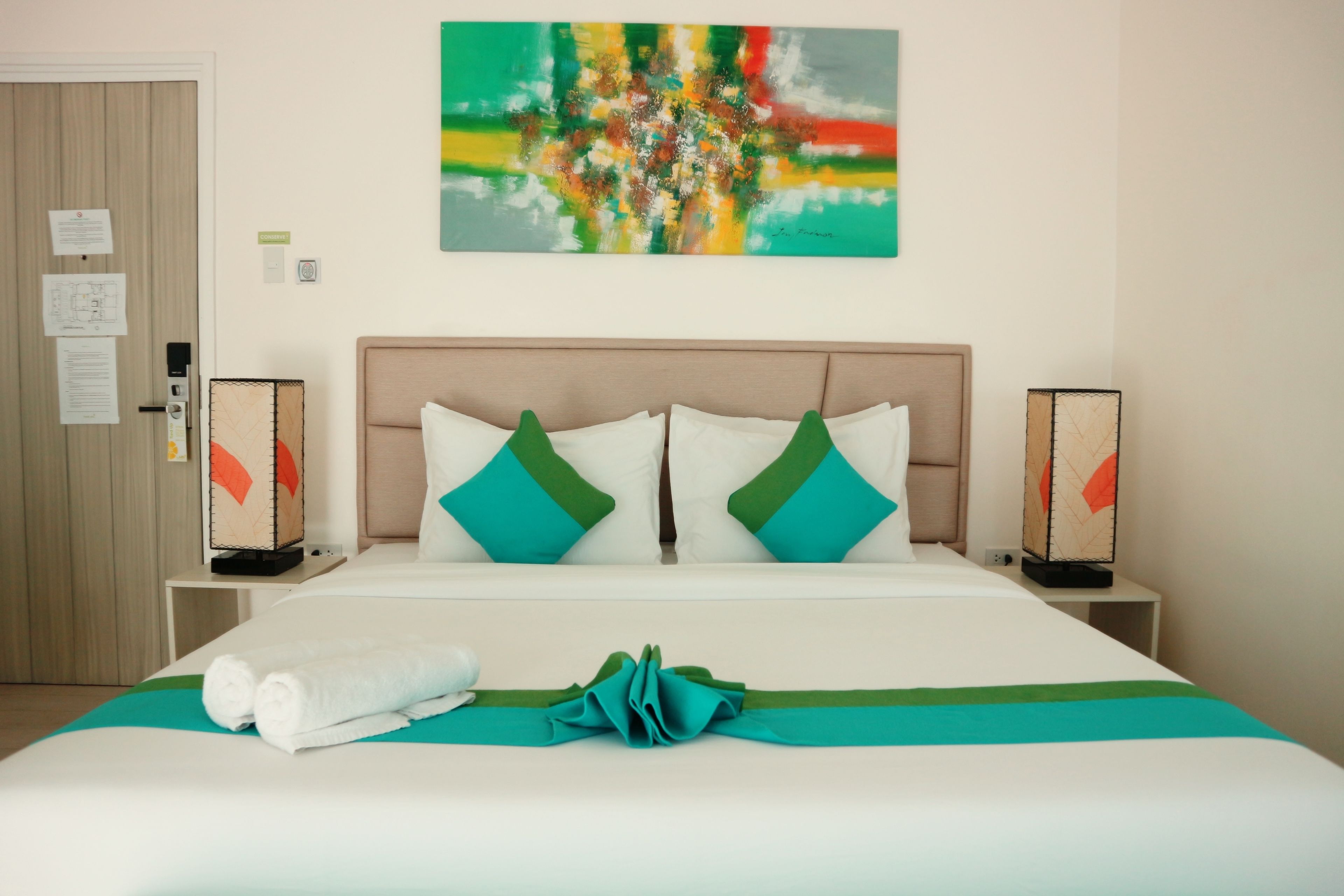 A hotel room with a bed, a headboard, two bedside tables, a wall art piece, and a door with a lock and a notice.