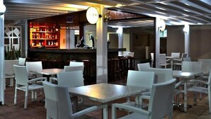 En restaurant med hvide fletmøbler, et barområde med hylder fyldt med flasker, og et ur på væggen.