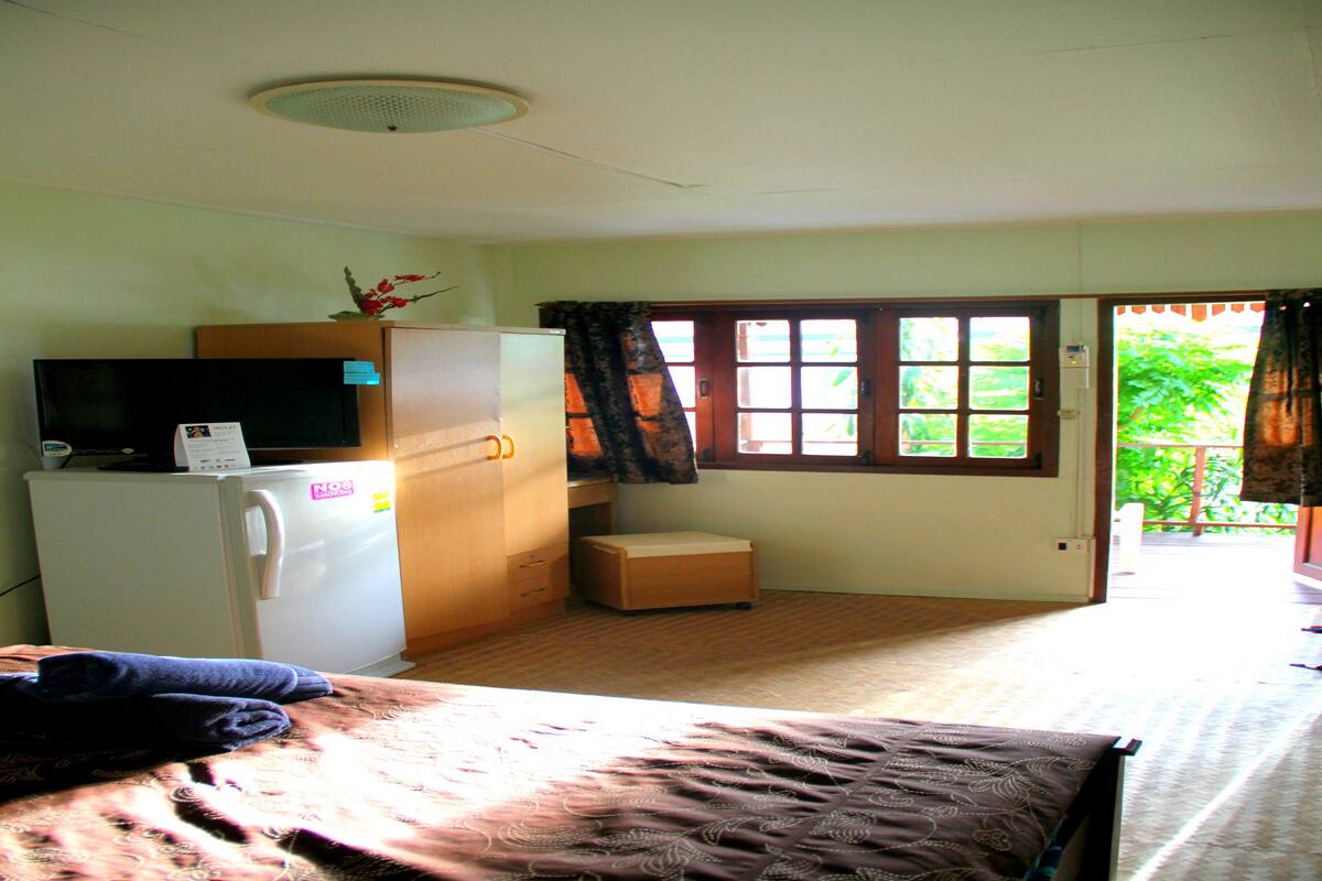 A compact room with a bed, a small wooden cabinet, a refrigerator, a television on top of the cabinet, and a window with curtains.