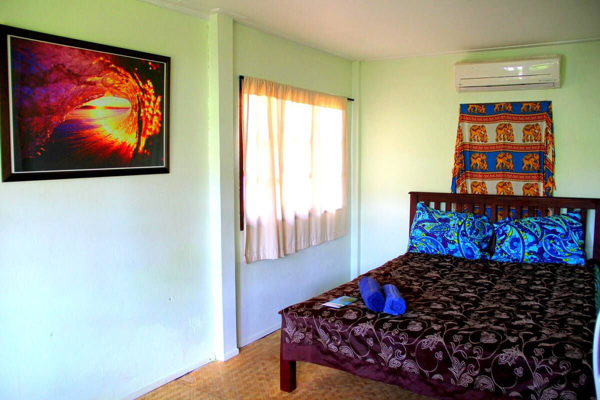 A bedroom with a bed, a window with curtains, an air conditioner, and a tapestry on the wall.