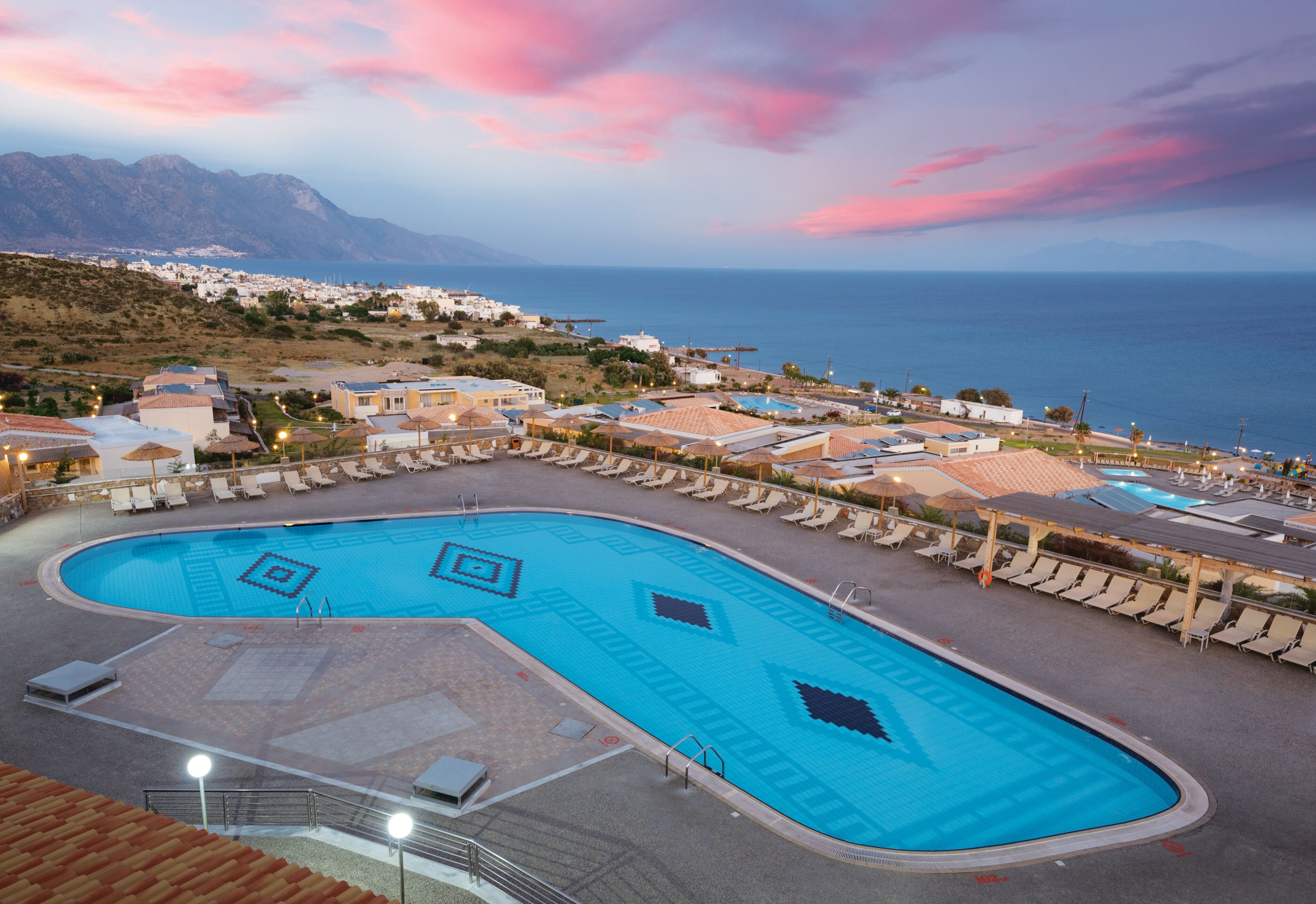 A swimming pool with geometric patterns, surrounded by lounge chairs and umbrellas, with a view of the sea and mountains in the background.