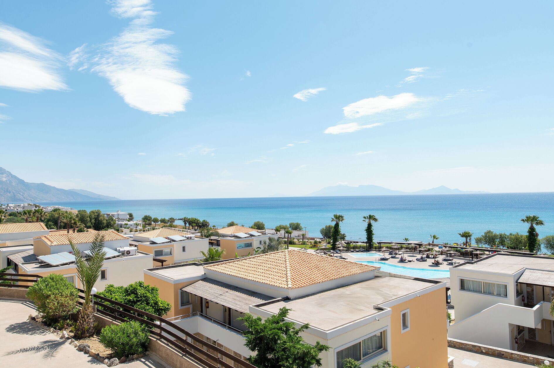 A coastal resort with multiple buildings, a swimming pool, and a view of the sea and mountains.