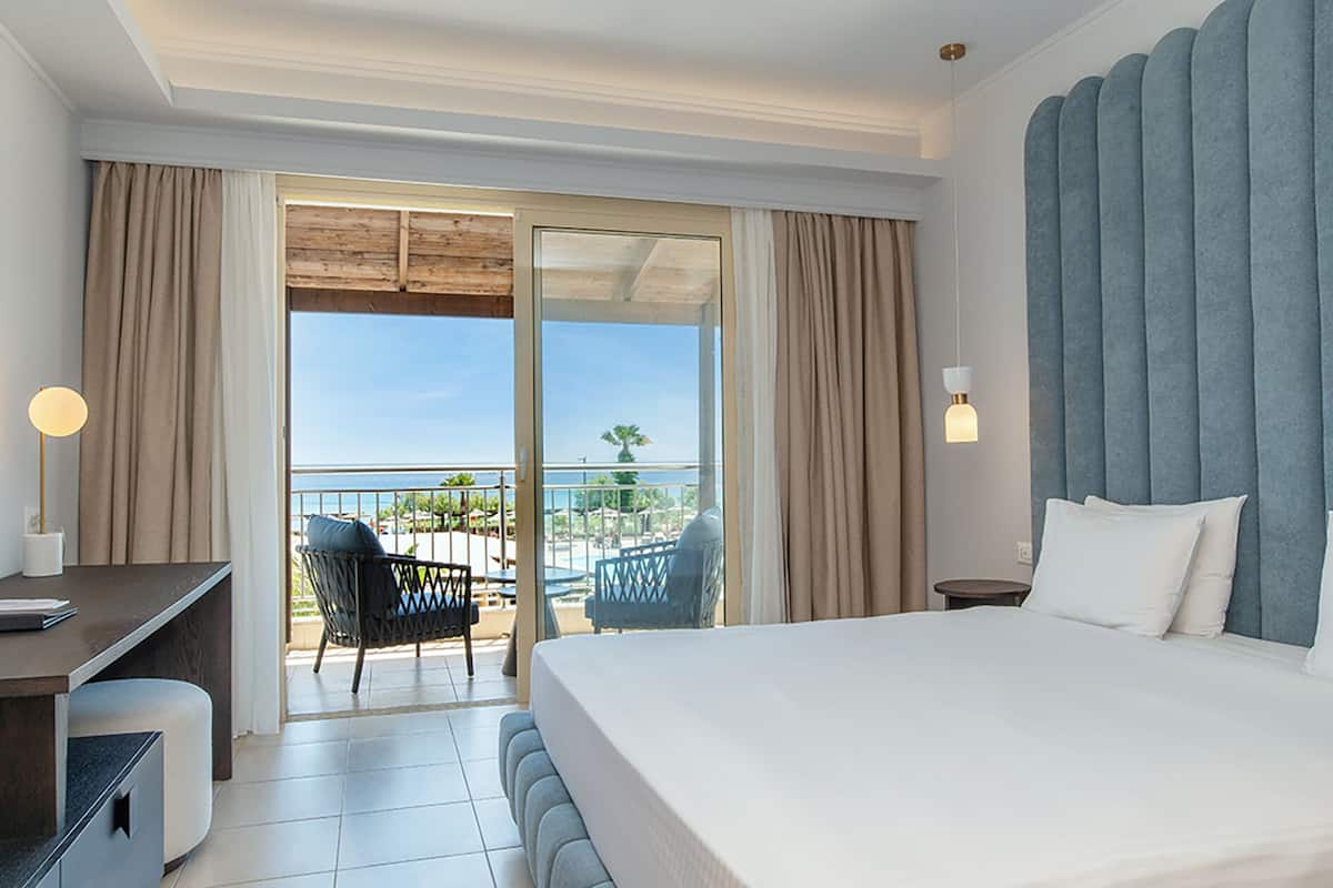 A hotel room with a bed, a desk, a chair, and a balcony with a view of the beach.