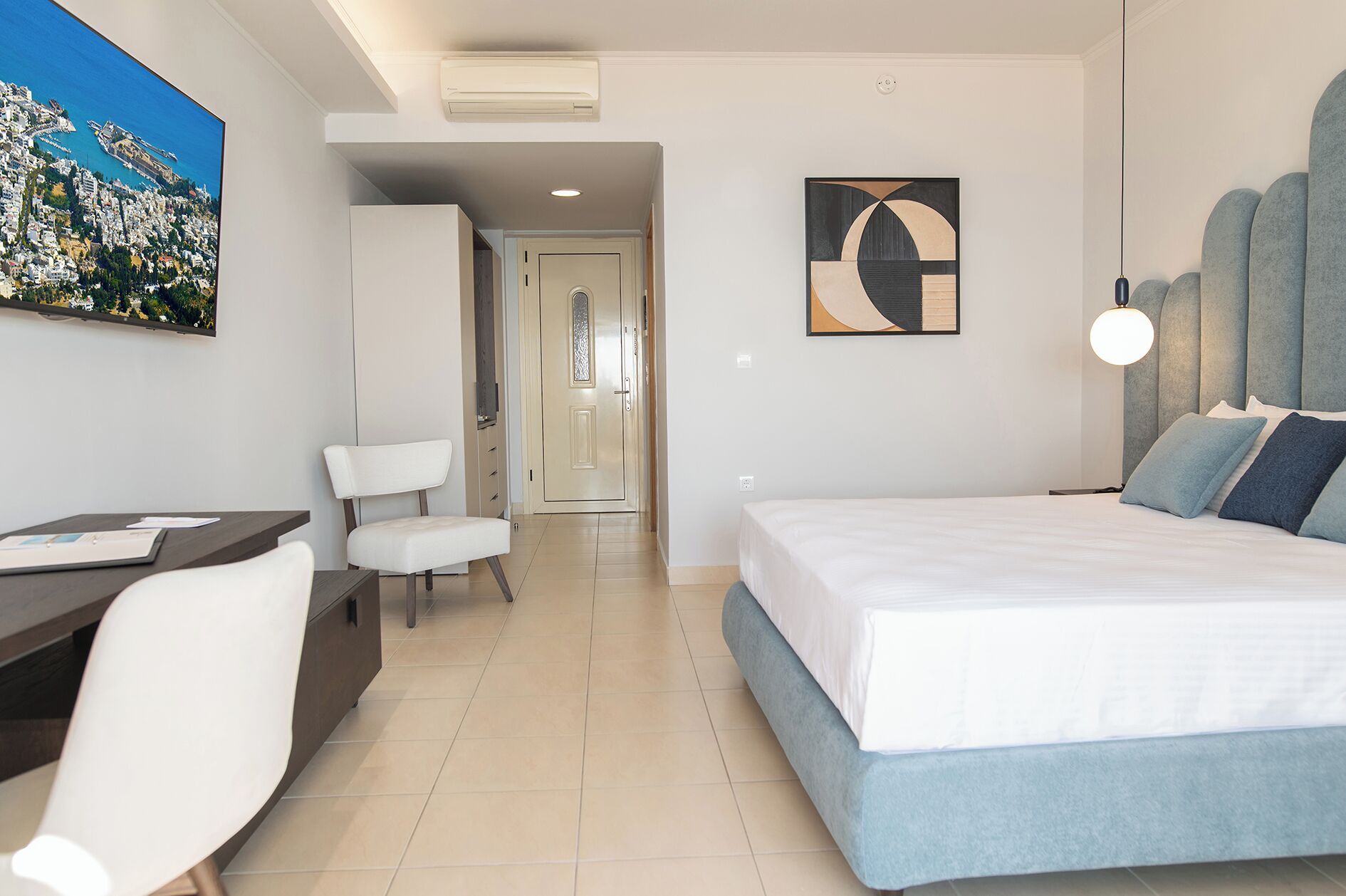 A modern hotel room with a bed, a desk, and a chair. There is a painting on the wall and a ceiling fan.