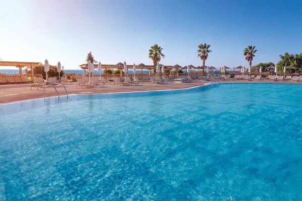 Una piscina azul cristalina con sillas de descanso y sombrillas, rodeada de palmeras y una terraza de madera.