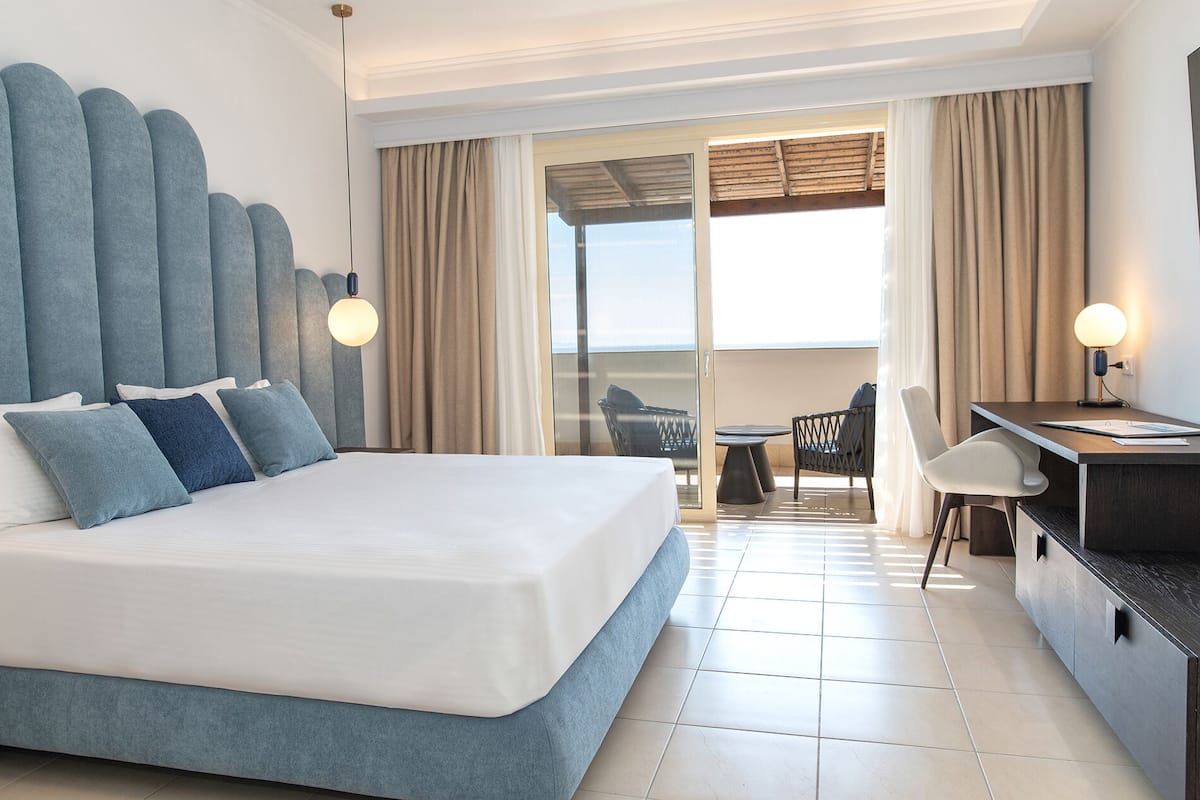 A hotel room with a large bed, a desk, and a view of the sea through a sliding glass door.