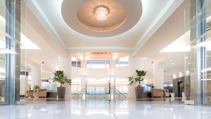 A spacious lobby with a central staircase, large windows, and potted plants. - Grand Blue Beach Hotel (Kos)