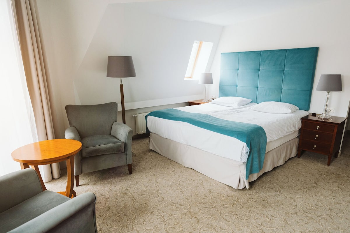 A hotel room with a large bed, two bedside tables, a chair, a small table, and a window.