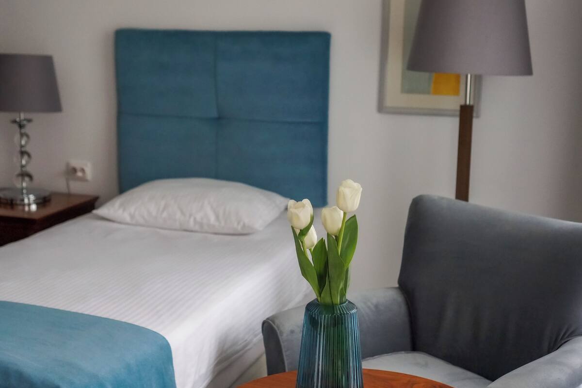 A hotel room with a bed, a blue headboard, a grey armchair, and a vase with white flowers.