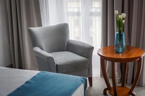A hotel room with a grey armchair, a round wooden table with a vase of flowers, and a bed with a blue pillow. - Hotel Śródka (Poznan)