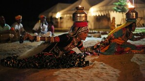 A group of people in traditional attire sitting on the ground, with a tent and a lit candle in the background. - Pushkar Adventure Desert Camp (Peesangan)