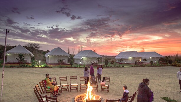 A group of people seated around a campfire in a sandy area with tents in the background. - Pushkar Adventure Desert Camp (Peesangan)