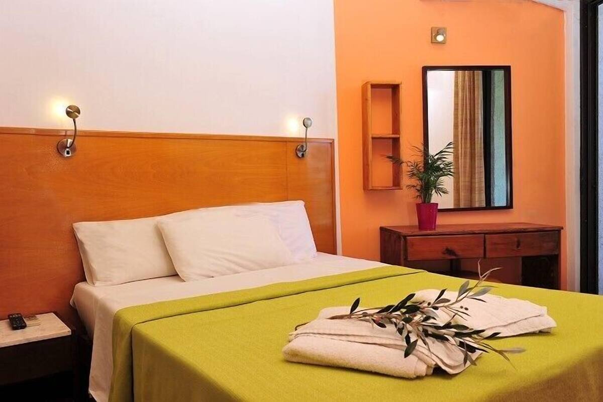 A hotel room with a bed, a nightstand, a bedside table, a mirror, a potted plant, and a window.