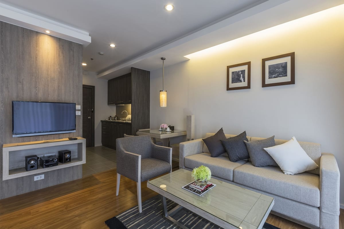 Een moderne woonkamer met een flatscreen televisie, een grijze bank, een salontafel en een eethoek op de achtergrond.