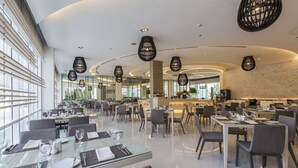 Een modern restaurant met glazen tafels, grijze stoelen en zwarte hanglampen.