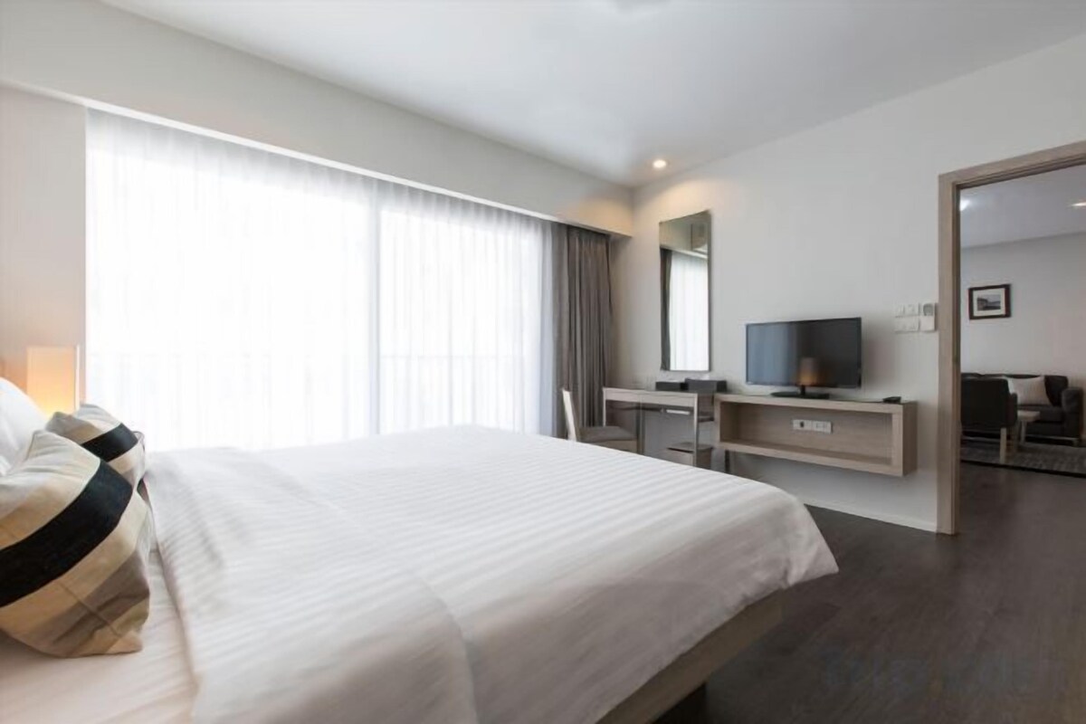 A hotel room with a large bed, a desk with a computer, a television, and a view of the outside through the window.