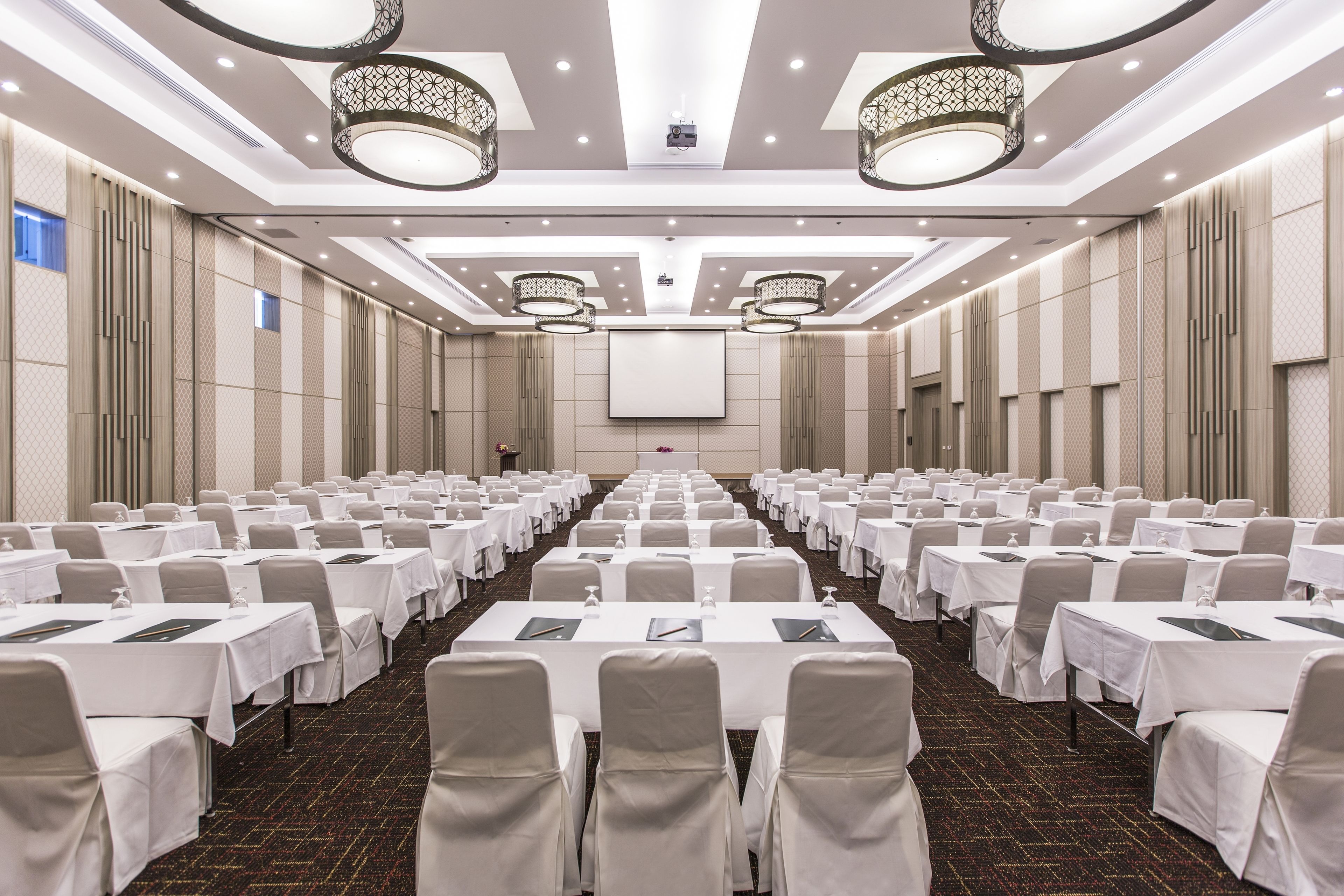 A conference room with rows of tables and chairs, a large screen at the front, and decorative lighting fixtures.