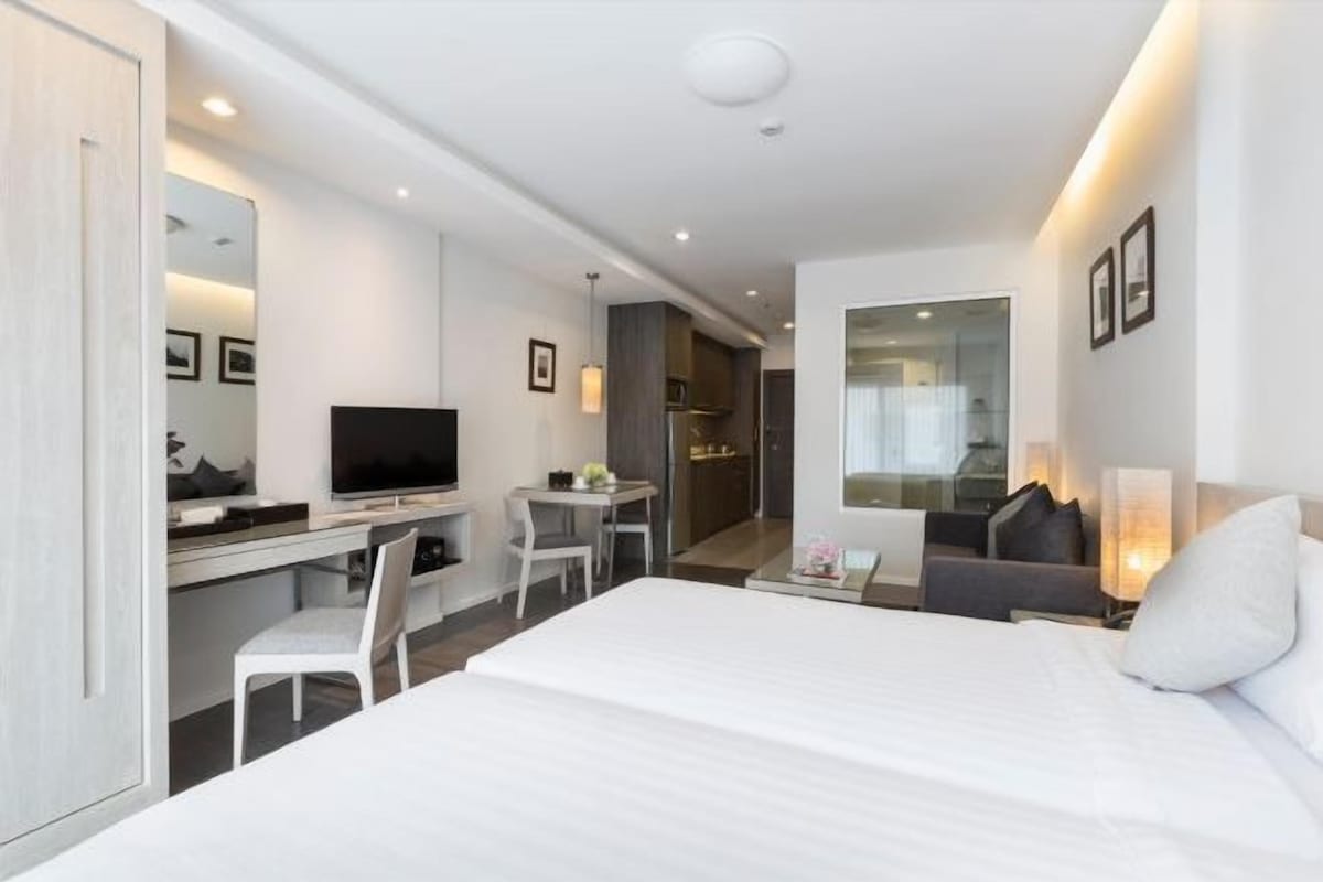 Una habitación de hotel moderna con una cama grande, un escritorio con televisor, una mesita y una zona de estar con sofá y sillones.