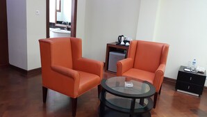 Una habitación con dos sillones anaranjados, una mesa de centro de vidrio, un refrigerador pequeño, un escritorio con un teléfono y una botella de agua, y un armario.