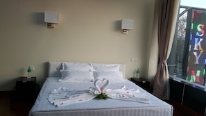 A hotel room with a neatly made bed, two bedside tables with water bottles, a decorative swan towel arrangement, and a large window with a colorful view. - Sky Man Hotel (Yangon)