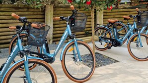 Tres bicicletas estacionadas en fila, cada una con una cesta y una etiqueta que dice 'Location Hotel Les Pieux'.