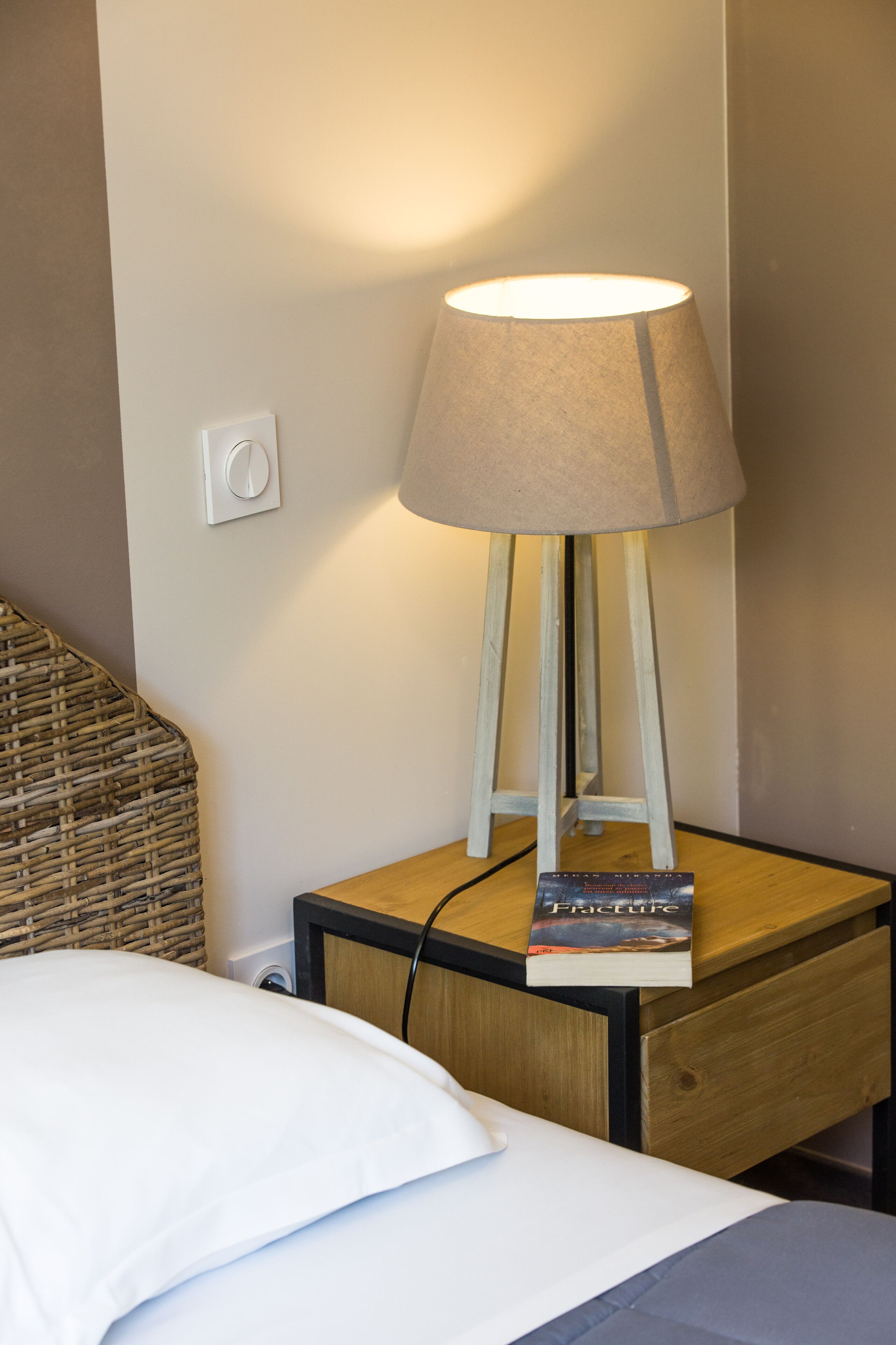 A bedside table with a lamp, a book titled "Fracture," and a power outlet.