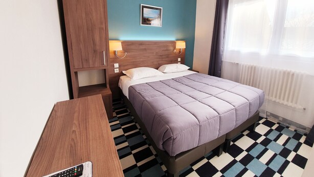 Una habitación de hotel con una cama, un armario de madera, un escritorio con un control remoto, piso a cuadros y una ventana con cortinas.