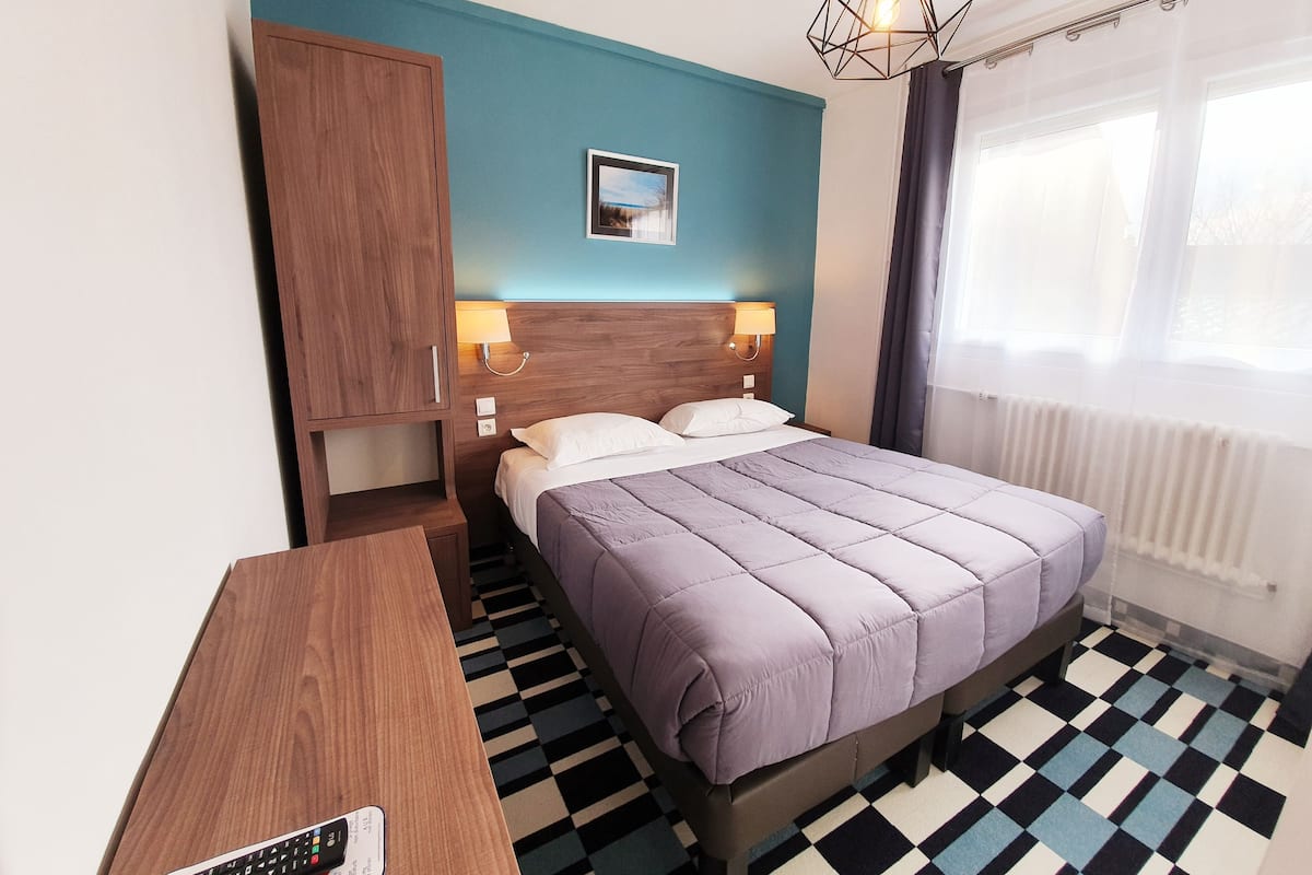 Hotellihuone, jossa on sänky, puinen vaatekaappi, kaukosäätimellä varustettu työpöytä, ruutukuviollinen lattia ja ikkuna, jossa on verhot.