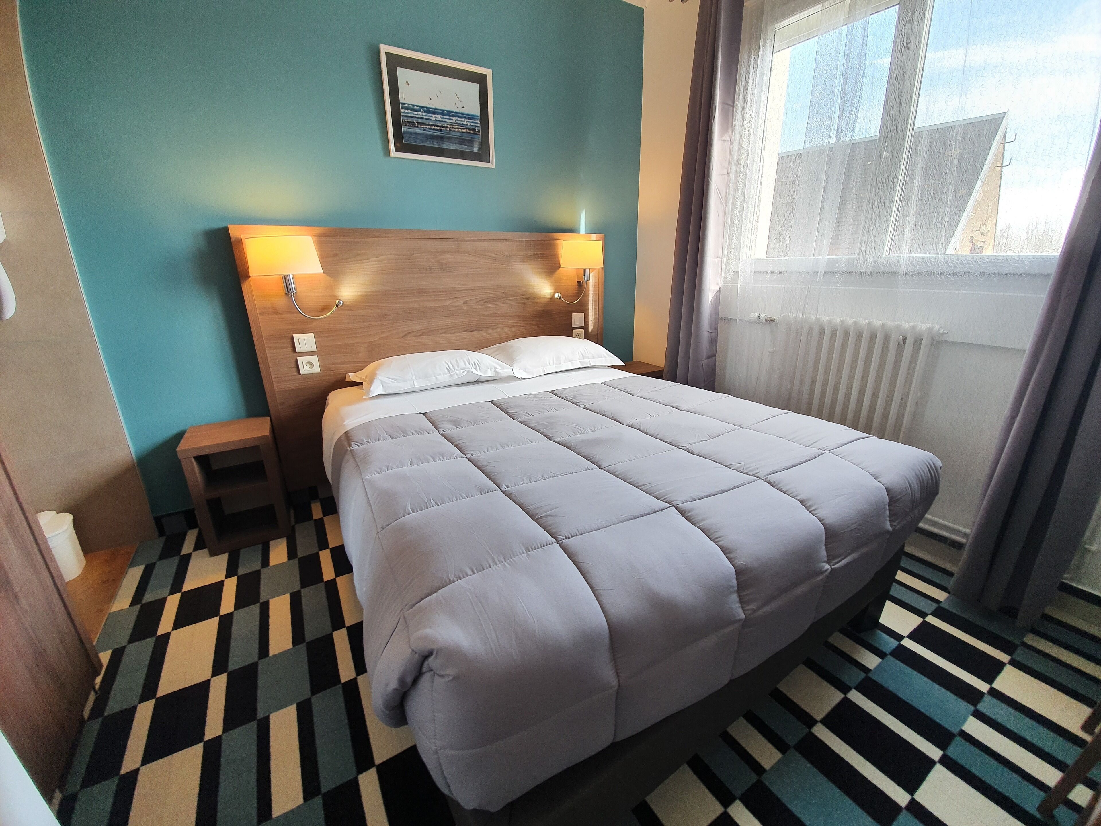 A hotel room with a checkered floor, a bed with a grey duvet, a headboard with wall-mounted lamps, and a window with curtains.