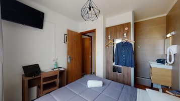 Une chambre d’hôtel avec un lit, un bureau avec un ordinateur portable, un manteau accroché et une salle de bain avec un miroir et un lavabo.