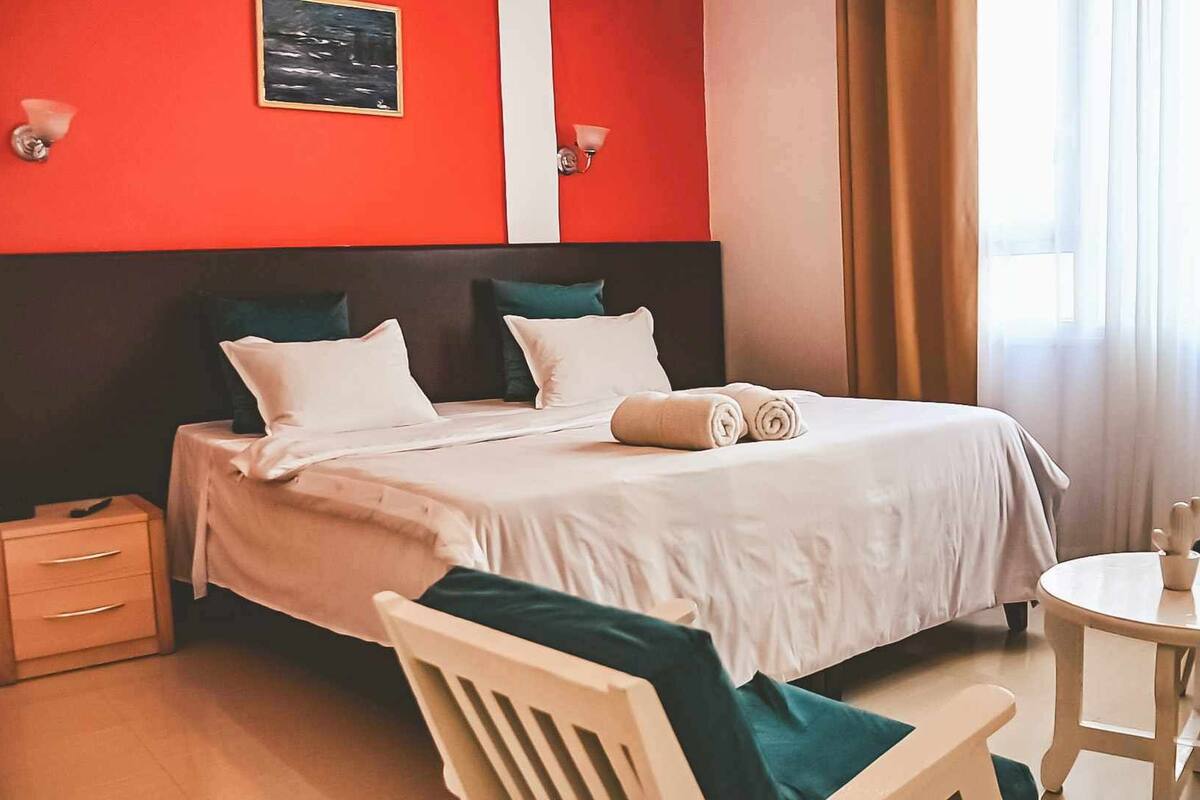 Een hotelkamer met een bed, nachtkastjes, een stoel, een klein rond tafeltje en een raam met gordijnen.