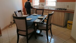 Een keuken met een eettafel voor vier personen, iemand kookt op het fornuis, en een raam met jaloezieën.