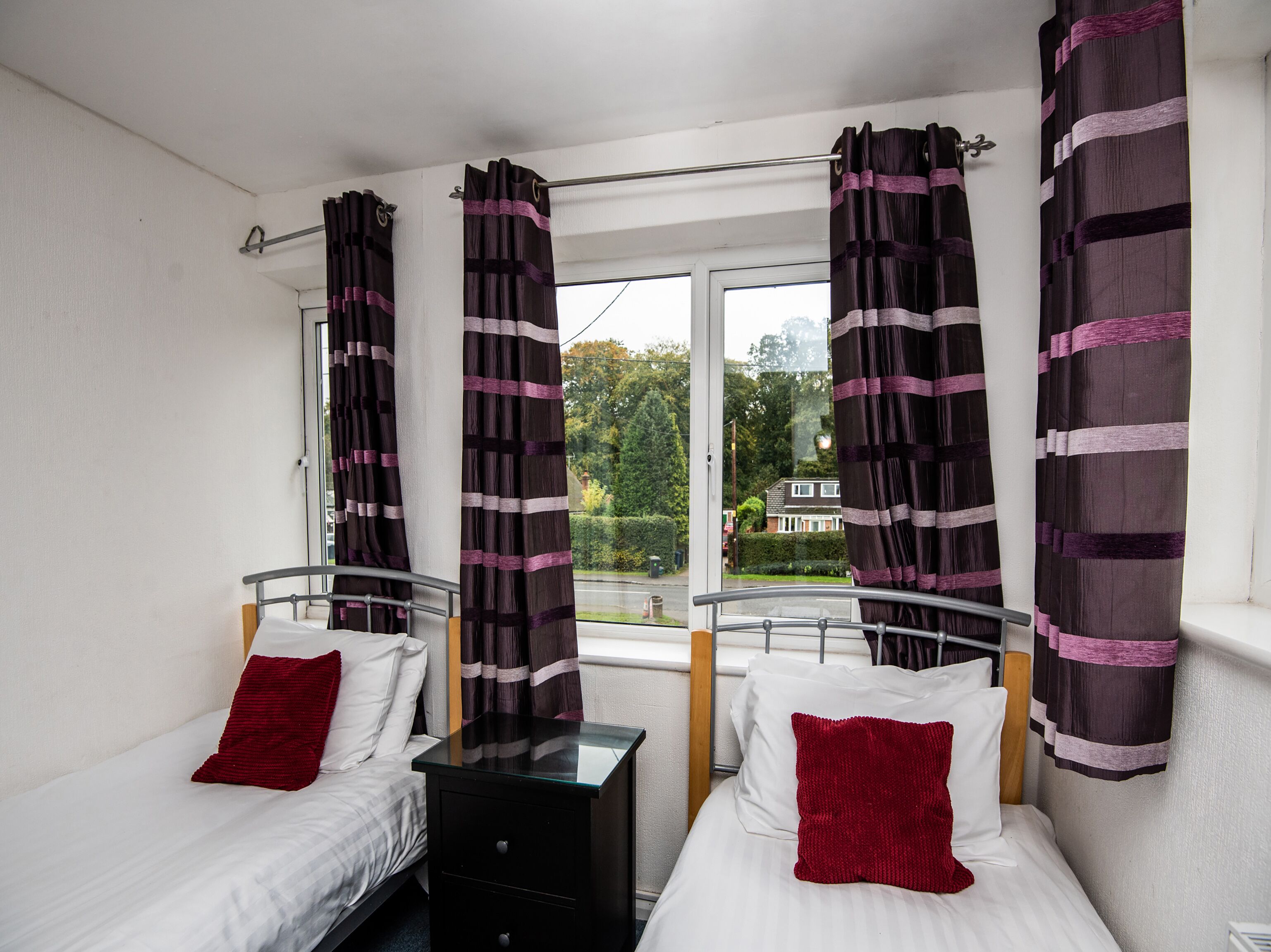 Habitación doble con dos camas, cada una con un almohadón rojo, y una ventana con cortinas.