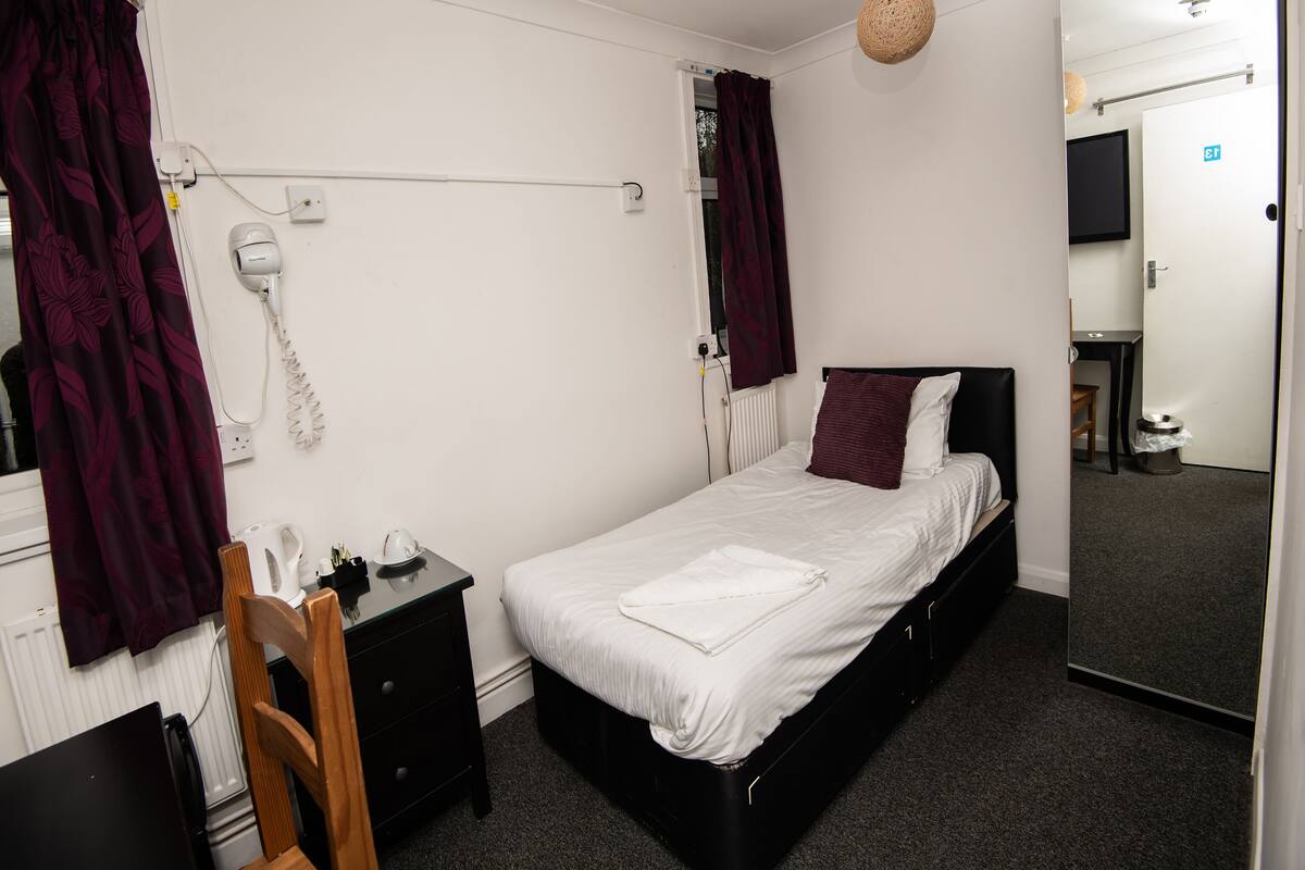 A bedroom with a single bed, a bedside table, a chair, a desk, a mirror, and a hanging light fixture.