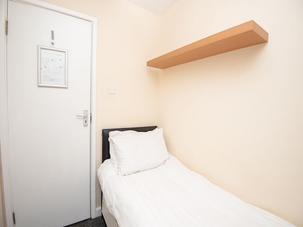 Una habitación pequeña y limpia con una cama individual, una puerta blanca y una estantería en la pared.