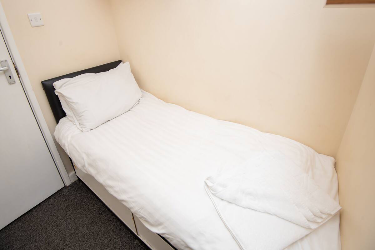 A single bed with white bedding in a small room with a door and a light switch on the wall.