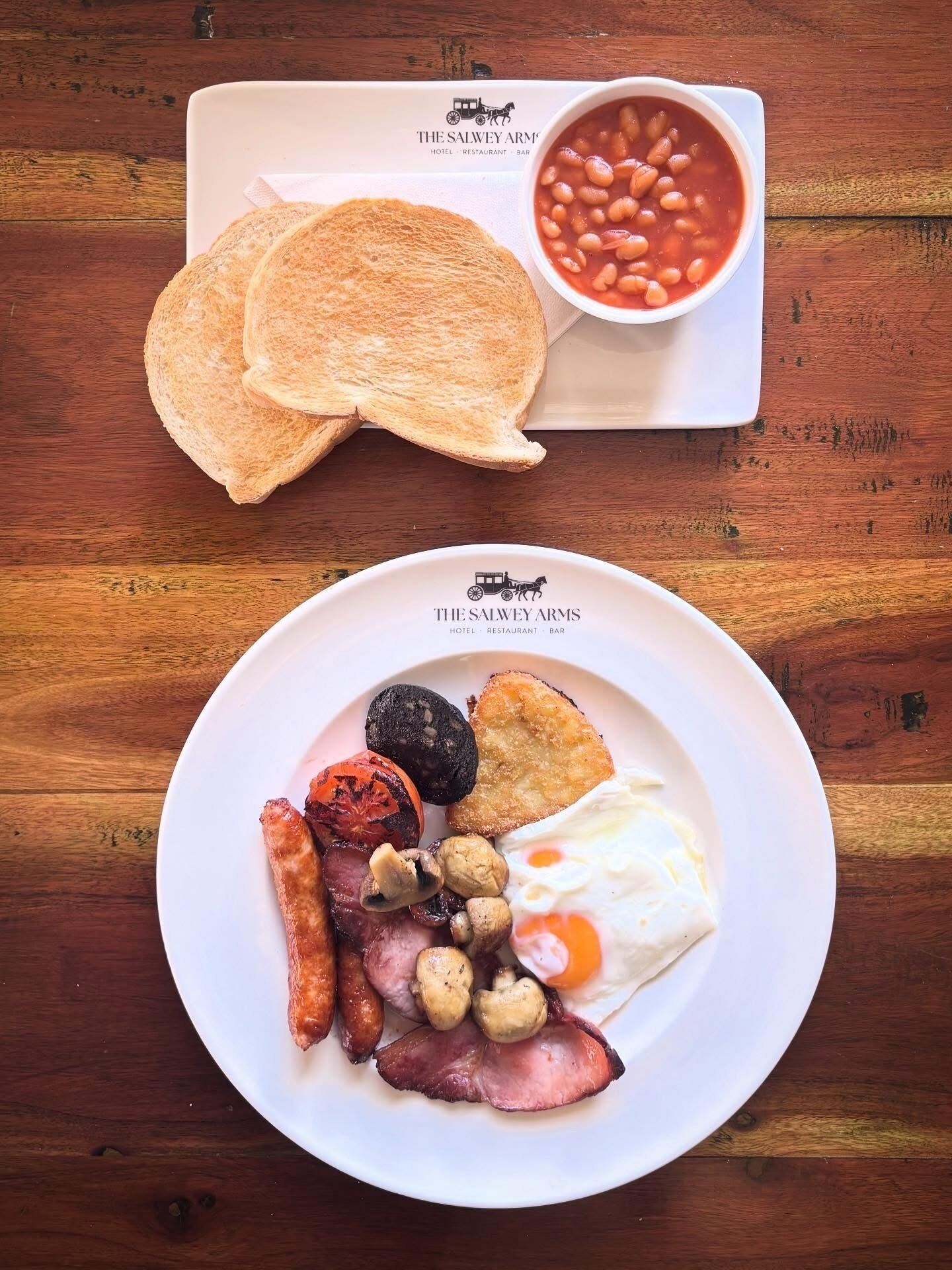 Une assiette de petit-déjeuner comprenant des œufs, des saucisses, des champignons et des toasts, accompagnée d’une portion de haricots cuits.