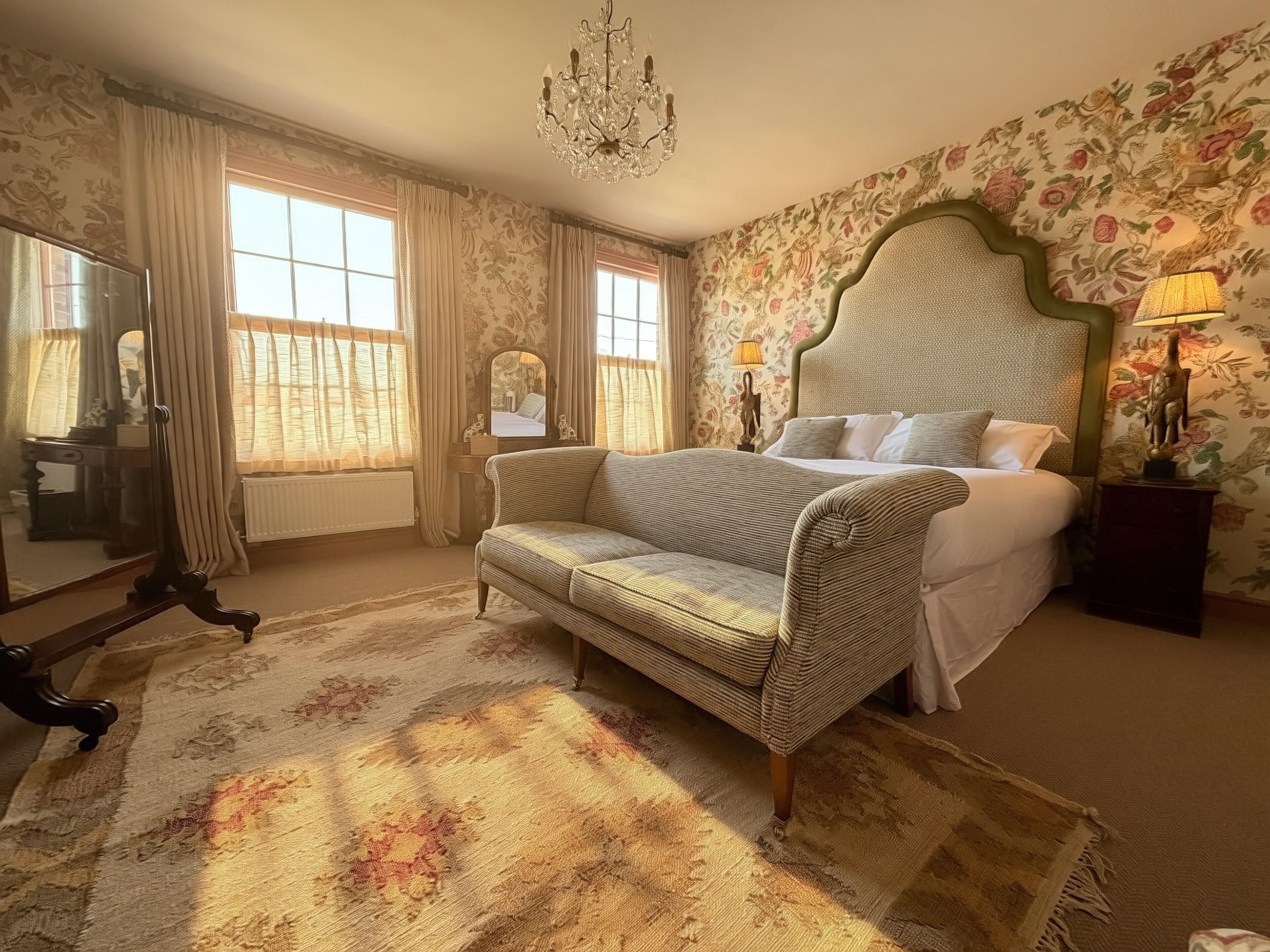 A bedroom with a large bed, a sofa, a mirror, a chandelier, and floral wallpaper.