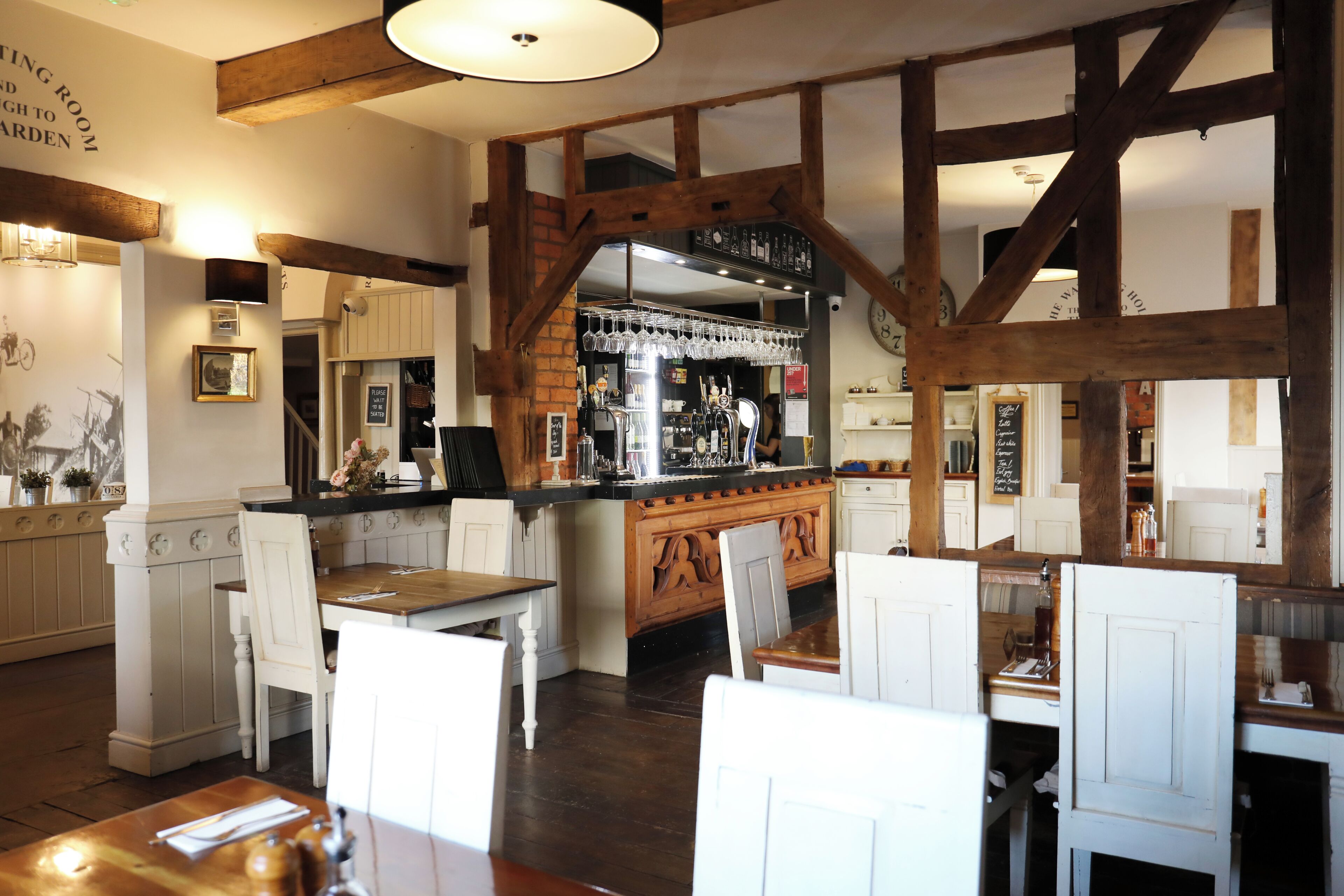 A restaurant with wooden beams, white chairs, a bar area, and a clock on the wall.