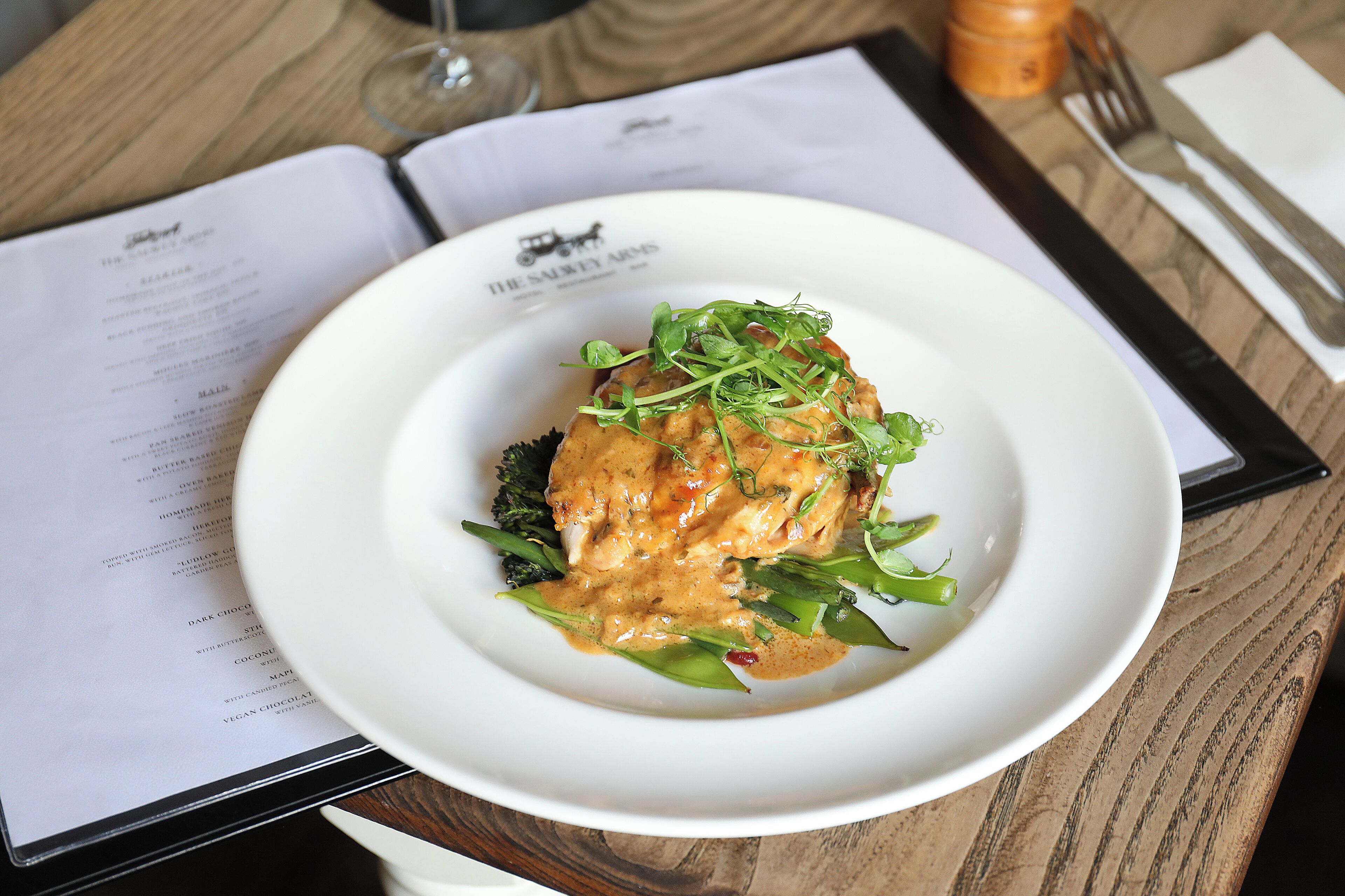 A white plate with a dish on a wooden table, menu, and cutlery.