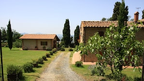Un chemin de gravier mène à une maison rustique à toit de tuiles, entourée de verdure et d’arbres.