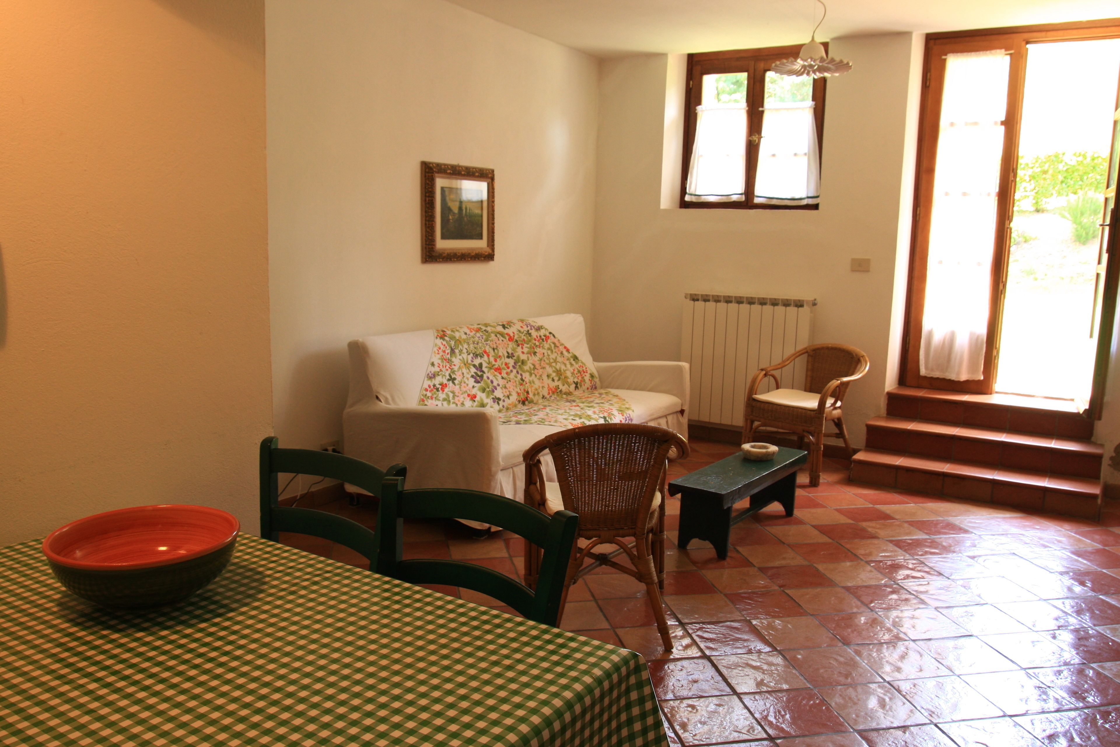 A room with a checkered table, a sofa with a floral cover, a wicker chair, a small table, and a window with a view of greenery.